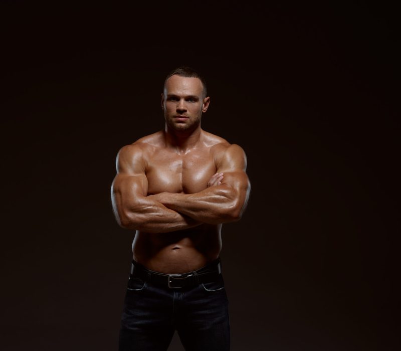 Male muscular athlete shows his power in studio, dark background. One man with athletic build, shirtless sportsman in jeans pants, active healthy lifestyle